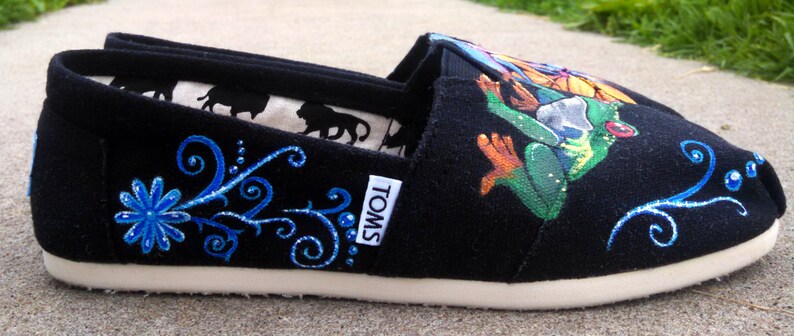 Puede incluir: Zapato negro sin cordones con suela blanca. El zapato presenta un dise&ntilde;o floral azul y una colorida ilustraci&oacute;n de una rana. El forro interior tiene un patr&oacute;n de silueta repetido. La marca "TOMS" es visible en el lateral.