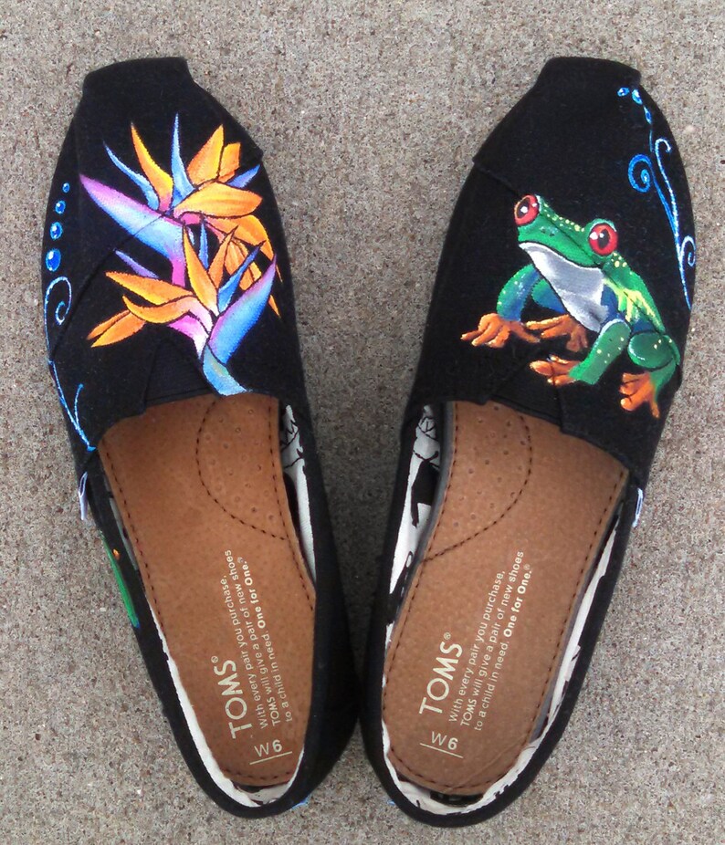 Puede incluir: Par de zapatos sin cordones TOMS negros, talla W6, con dise&ntilde;os pintados a mano. Un zapato presenta una colorida flor de ave del para&iacute;so, el otro una rana arb&oacute;rea verde. Ambos tienen plantillas marrones y el logo TOMS.