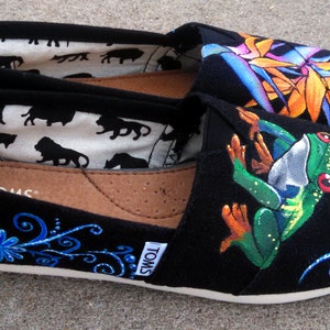 Puede incluir: Zapatillas negras sin cordones con dise&ntilde;os pintados coloridos. Un zapato presenta una rana verde con ojos rojos, el otro una flor tropical. Los zapatos tienen suelas blancas y la marca "TOMS" es visible.