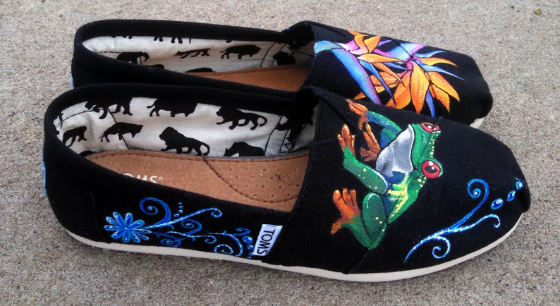 Puede incluir: Zapatillas negras sin cordones con dise&ntilde;os pintados coloridos. Un zapato presenta una rana verde con ojos rojos, el otro una flor tropical. Los zapatos tienen suelas blancas y la marca "TOMS" es visible.