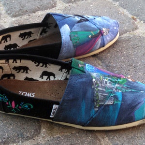 K&ouml;nnte beinhalten: Ein Paar Slip-on-Leinenschuhe mit einem farbenfrohen, bemalten Design. Die Schuhe haben eine schwarze Spitze und Ferse, mit einem gemusterten Innenraum und einem bemalten Design auf der Oberseite. Das Design umfasst Blau-, Gr&uuml;n- und Lilat&ouml;ne.