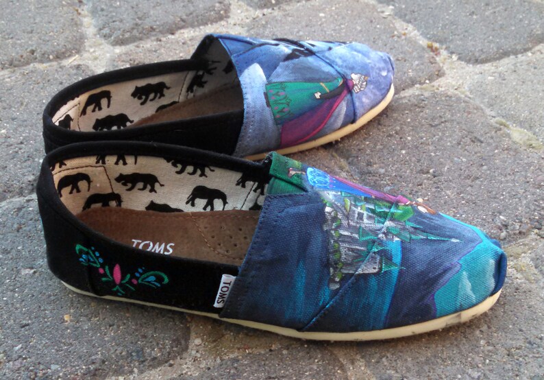 K&ouml;nnte beinhalten: Ein Paar Slip-on-Leinenschuhe mit einem farbenfrohen, bemalten Design. Die Schuhe haben eine schwarze Spitze und Ferse, mit einem gemusterten Innenraum und einem bemalten Design auf der Oberseite. Das Design umfasst Blau-, Gr&uuml;n- und Lilat&ouml;ne.
