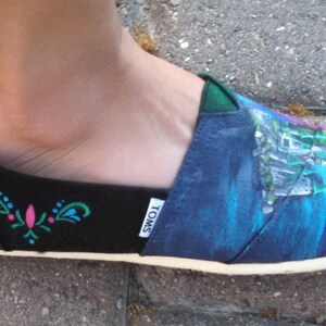 K&ouml;nnte beinhalten: Schwarzer und blauer Slip-On-Schuh mit wei&szlig;er Sohle. Der Schuh hat ein gemaltes Design eines Schlosses und ein Blumenmuster. Der Markenname "TOMS" ist auf der Seite des Schuhs sichtbar. Der Schuh wird an einem Fu&szlig; getragen.