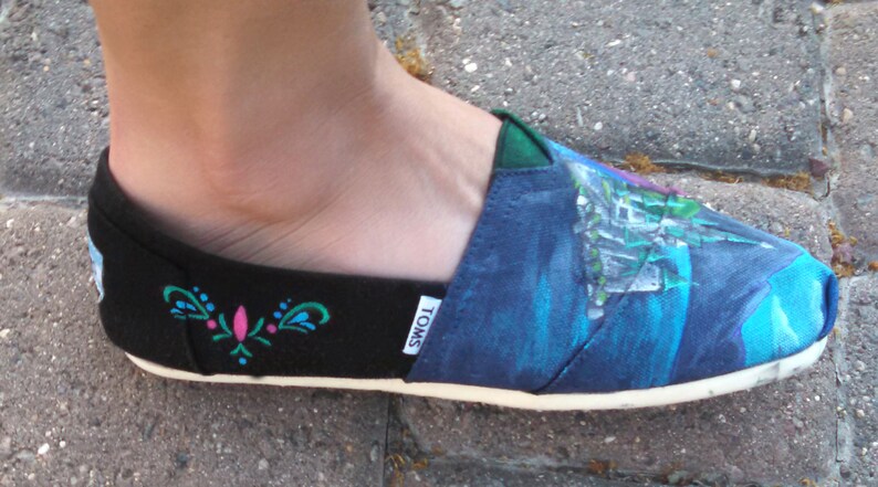 K&ouml;nnte beinhalten: Schwarzer und blauer Slip-On-Schuh mit wei&szlig;er Sohle. Der Schuh hat ein gemaltes Design eines Schlosses und ein Blumenmuster. Der Markenname "TOMS" ist auf der Seite des Schuhs sichtbar. Der Schuh wird an einem Fu&szlig; getragen.