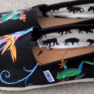 Puede incluir: Un par de zapatos negros de lona sin cordones con dise&ntilde;os pintados a mano. Uno presenta una rana verde, el otro una flor tropical. El forro interior tiene un patr&oacute;n repetido de siluetas de animales negros.
