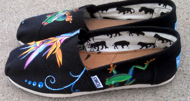 Puede incluir: Un par de zapatos negros de lona sin cordones con dise&ntilde;os pintados a mano. Uno presenta una rana verde, el otro una flor tropical. El forro interior tiene un patr&oacute;n repetido de siluetas de animales negros.