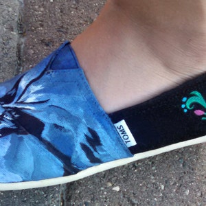 K&ouml;nnte beinhalten: Blauer und schwarzer Slip-On-Schuh mit floralem Design. Die Spitze des Schuhs weist ein blaues Blumenmuster auf, w&auml;hrend die Ferse schwarz ist mit einem dekorativen Design in T&uuml;rkis und Rosa. Der Schuh hat eine wei&szlig;e Sohle und die Marke "TOMS" ist sichtbar.