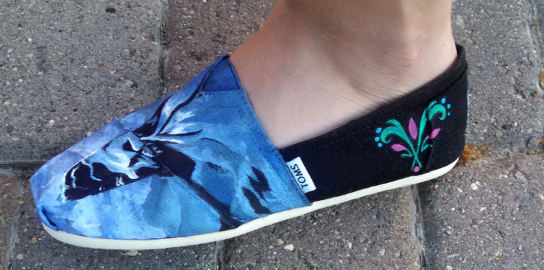 K&ouml;nnte beinhalten: Blauer und schwarzer Slip-On-Schuh mit floralem Design. Die Spitze des Schuhs weist ein blaues Blumenmuster auf, w&auml;hrend die Ferse schwarz ist mit einem dekorativen Design in T&uuml;rkis und Rosa. Der Schuh hat eine wei&szlig;e Sohle und die Marke "TOMS" ist sichtbar.