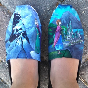 K&ouml;nnte beinhalten: Ein Paar Slipper mit blauem und gr&uuml;nem Design, das Charaktere und Szenen aus einem beliebten Animationsfilm zeigt. Die Schuhe haben schwarze Sohlen und die Oberteile sind mit Bildern eines Schlosses, Bergen und Charakteren verziert.