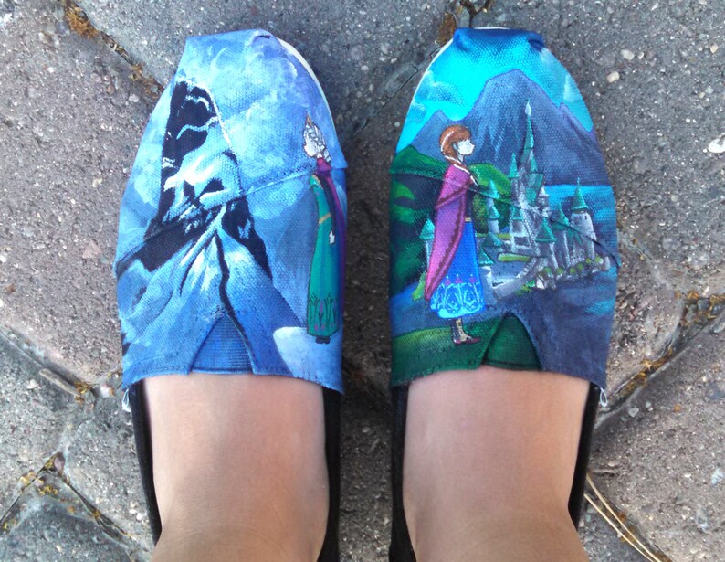 K&ouml;nnte beinhalten: Ein Paar Slipper mit blauem und gr&uuml;nem Design, das Charaktere und Szenen aus einem beliebten Animationsfilm zeigt. Die Schuhe haben schwarze Sohlen und die Oberteile sind mit Bildern eines Schlosses, Bergen und Charakteren verziert.