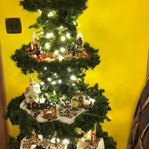 Puede incluir: Un &aacute;rbol de Navidad de varios niveles decorado con casas en miniatura y luces. El &aacute;rbol est&aacute; decorado con material blanco similar a la nieve y est&aacute; coronado con una iglesia en miniatura. El &aacute;rbol est&aacute; frente a una pared amarilla con la palabra "JOY" en luces.