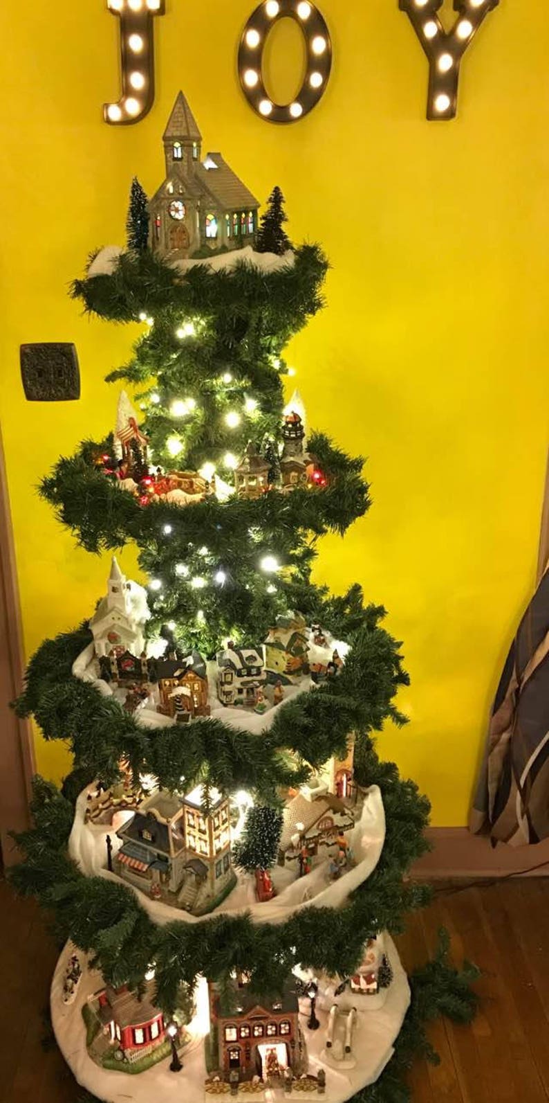 Puede incluir: Un &aacute;rbol de Navidad de varios niveles decorado con casas en miniatura y luces. El &aacute;rbol est&aacute; decorado con material blanco similar a la nieve y est&aacute; coronado con una iglesia en miniatura. El &aacute;rbol est&aacute; frente a una pared amarilla con la palabra "JOY" en luces.