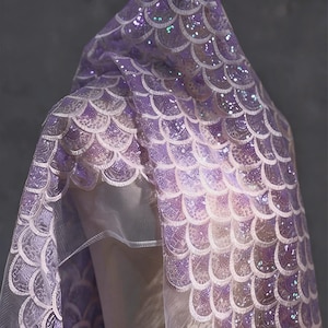 May include: A lavender-colored, hooded cape with a scale-like pattern. The fabric is embellished with sequins, creating a shimmering effect. The cape is draped over a mannequin, showcasing its design and texture.
