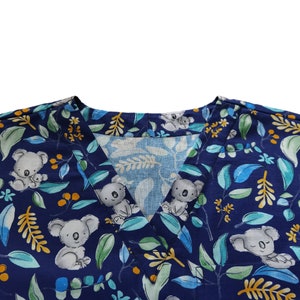 May include: Navy blue V-neck scrub top featuring a repeating pattern of koalas, leaves, and berries. The koalas are grey and white, and the leaves are green and blue. The berries are yellow. The fabric is a soft, lightweight material.