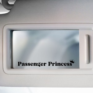 Op de afbeelding: Een witte autozonneklep met een spiegel en een zwarte sticker met de tekst "Passenger Princess" met een kroon.