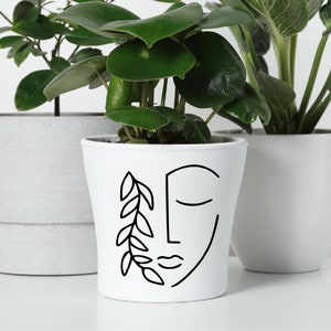 May include: White ceramic planter with a black line drawing of a face and a leafy branch. The planter is filled with a green plant.