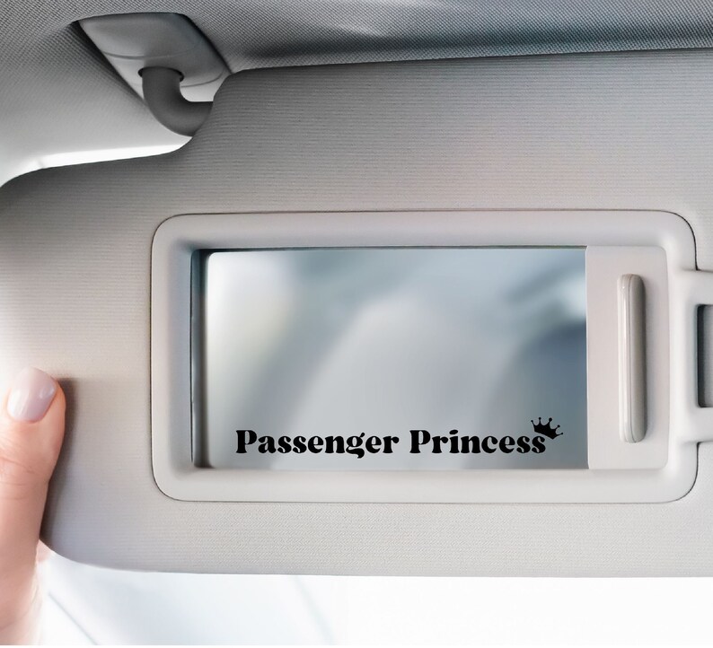 Op de afbeelding: Een witte autozonneklep met een spiegel en een zwarte sticker met de tekst "Passenger Princess" met een kroon.