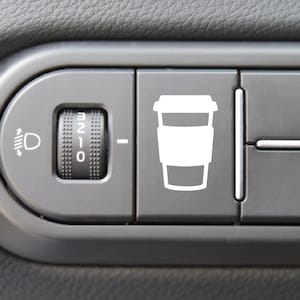 May include: Close-up of a car's dashboard with a white coffee cup icon on a button. The button is part of a series of buttons and a dial for the headlights. The dashboard is gray.