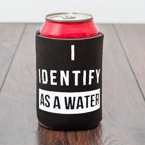 Op de afbeelding: Zwarte en witte blikjeskoeler met de tekst "I identify as a water" erop gedrukt. De koeler houdt een rood blikje vast.