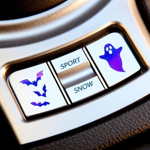 May include: A silver car console button panel featuring custom decals. The left button displays three purple and blue gradient bats, the center button shows the text SPORT and SNOW, and the right button features a purple and blue gradient ghost.