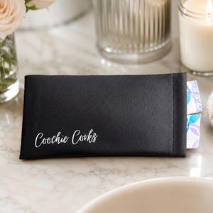 May include: A rectangular black faux leather pouch with a textured surface. The text "Coochie Corks" is printed in white script on the front. The pouch is slightly open on the right side, revealing two small, patterned, individually wrapped items.