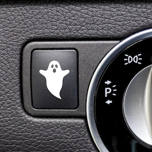 Könnte beinhalten: Nahaufnahme eines schwarzen Autoinnenraums. Eine schwarze Taste mit einem weißen Geistersymbol befindet sich neben einem kreisförmigen Zifferblatt mit Lichtsymbolen. Der Hintergrund ist eine strukturierte, dunkelgraue Oberfläche.