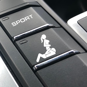 Peut inclure: Un bouton int&eacute;rieur de voiture noir avec un autocollant graphique blanc repr&eacute;sentant deux silhouettes dans une position sexuelle. Le bouton est situ&eacute; juste en dessous d'un bouton marqu&eacute; SPORT en texte blanc, sur une console en plastique noir.