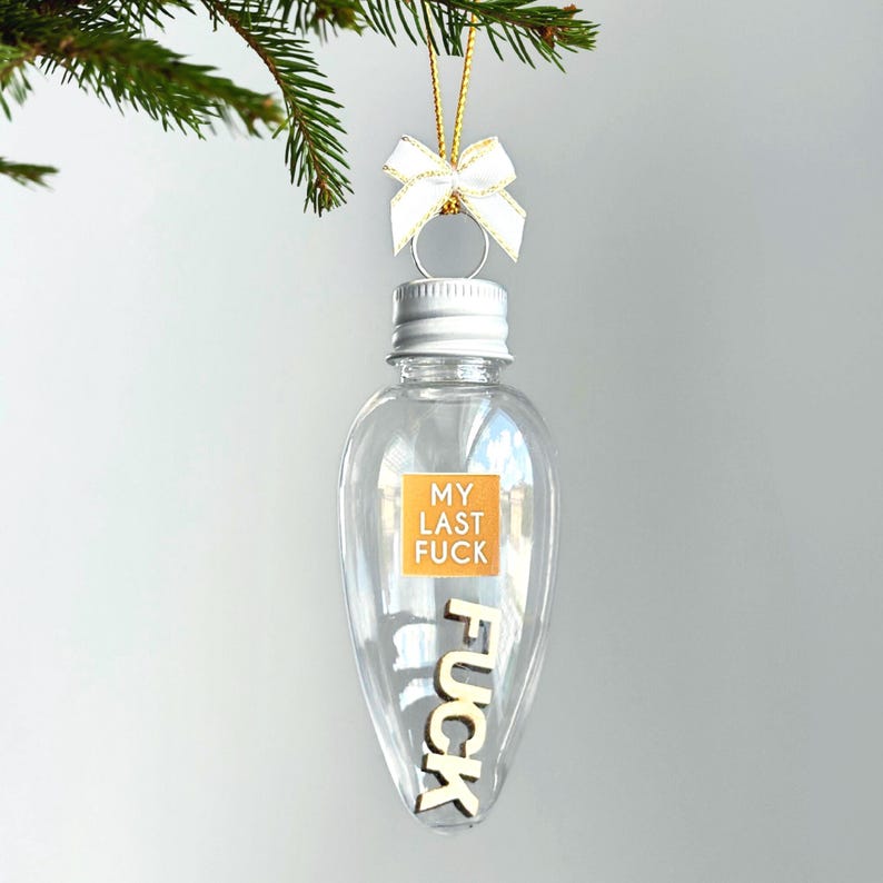 May include: Clear, lightbulb-shaped Christmas ornament with a silver cap and a white bow. The ornament has the words "MY LAST FUCK" on a small orange square and the word "FUCK" in wooden letters inside.
