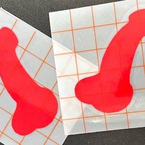 May include: Two red vinyl stickers, each featuring a stylized phallus shape. The stickers are on a clear backing with an orange grid pattern. The stickers are likely for decorative purposes.