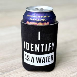 Op de afbeelding: Een zwarte neopreen blikjeskoeler met de tekst "I Identify as a Water" in wit. De koeler bevat een blikje Goose Island Beer Co. Neon bier.