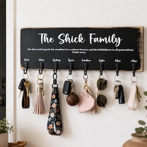 May include: A black wooden key holder with the text "The Shick Family" in white script. Below the text are the names of family members. Various keys and keychains hang from the hooks, including a floral strap and a pink heart-shaped keychain.
