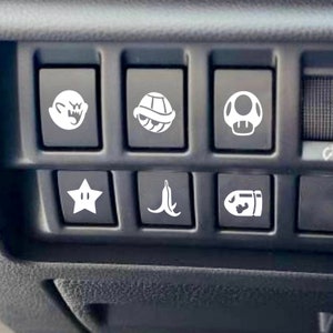 May include: A set of six black rectangular vehicle dashboard buttons, each featuring a white vinyl decal. The icons include a ghost, a turtle shell, a mushroom, a star, a banana peel, and a bullet, arranged in two rows of three within the console.