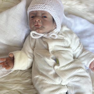 May include: A realistic reborn doll with dark hair, wearing a white knitted bonnet with a chin tie and a white long-sleeved ribbed onesie featuring a small star detail on the chest. The doll has detailed facial features and is lying down.