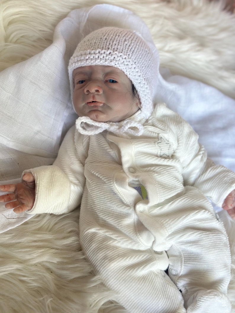 May include: A realistic reborn doll with dark hair, wearing a white knitted bonnet with a chin tie and a white long-sleeved ribbed onesie featuring a small star detail on the chest. The doll has detailed facial features and is lying down.