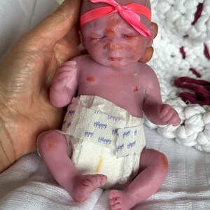 K&ouml;nnte beinhalten: Eine kleine, violett get&ouml;nte Silikonpuppe mit geschlossenen Augen und kahlem Kopf, die ein leuchtend neonpinkes Schleifenstirnband tr&auml;gt. Die Puppe ist mit einer wei&szlig;en Einwegwindel bekleidet, die ein wiederkehrendes blaues "Happy"-Textmuster aufweist.