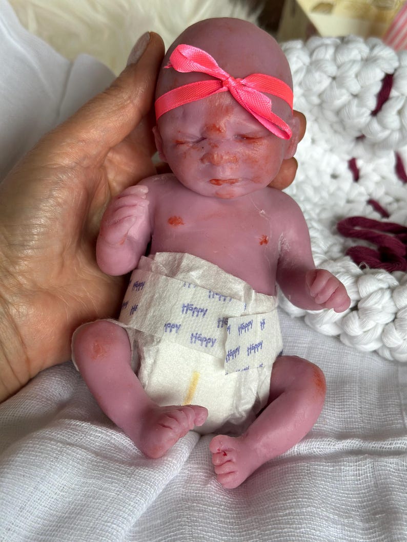K&ouml;nnte beinhalten: Eine kleine, violett get&ouml;nte Silikonpuppe mit geschlossenen Augen und kahlem Kopf, die ein leuchtend neonpinkes Schleifenstirnband tr&auml;gt. Die Puppe ist mit einer wei&szlig;en Einwegwindel bekleidet, die ein wiederkehrendes blaues "Happy"-Textmuster aufweist.