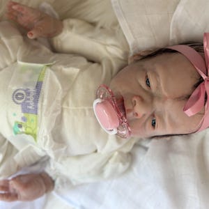 May include: A realistic reborn doll with dark hair, wearing a cream-colored long-sleeved onesie and a pink satin headband. The doll has a light skin tone, blue eyes, and a pink pacifier in its mouth. It is wearing a disposable diaper with green and purple graphics.