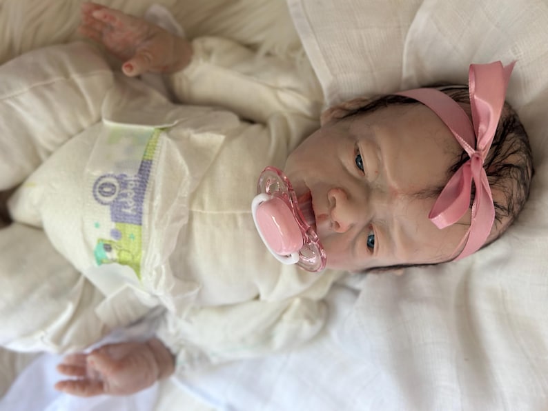 May include: A realistic reborn doll with dark hair, wearing a cream-colored long-sleeved onesie and a pink satin headband. The doll has a light skin tone, blue eyes, and a pink pacifier in its mouth. It is wearing a disposable diaper with green and purple graphics.