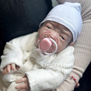 May include: A realistic baby doll with pale skin, blue eyes, and a light pink pacifier in its mouth. The doll wears a plain white beanie cap and a white long-sleeved textured cardigan with small buttons. The doll has detailed fingers and skin texture.