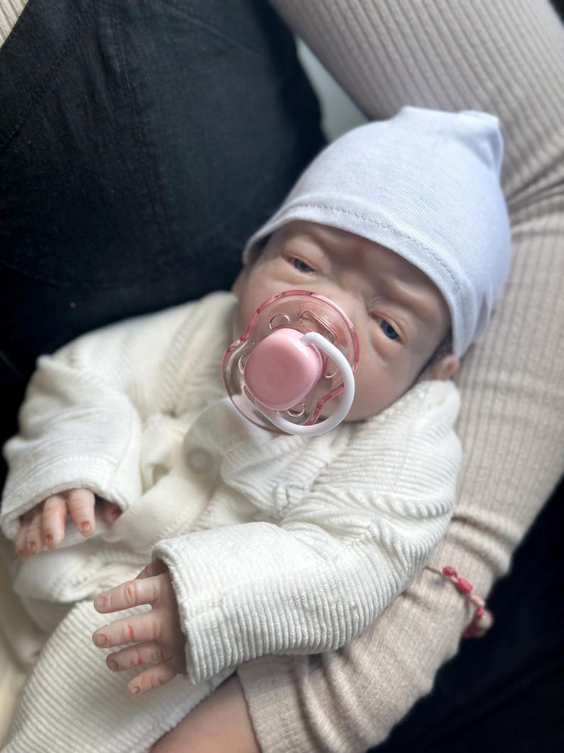 May include: A realistic baby doll with pale skin, blue eyes, and a light pink pacifier in its mouth. The doll wears a plain white beanie cap and a white long-sleeved textured cardigan with small buttons. The doll has detailed fingers and skin texture.