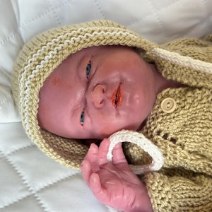 Puede incluir: Una mu&ntilde;eca beb&eacute; realista con un su&eacute;ter con capucha beige tejido y un gorro a juego con ribete blanco. La mu&ntilde;eca tiene un tono de piel realista, ojos azules y boca abierta. El su&eacute;ter tiene cierre de bot&oacute;n y el gorro tiene un cord&oacute;n blanco.