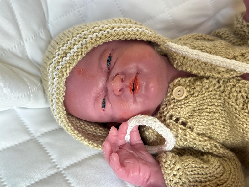 Puede incluir: Una mu&ntilde;eca beb&eacute; realista con un su&eacute;ter con capucha beige tejido y un gorro a juego con ribete blanco. La mu&ntilde;eca tiene un tono de piel realista, ojos azules y boca abierta. El su&eacute;ter tiene cierre de bot&oacute;n y el gorro tiene un cord&oacute;n blanco.