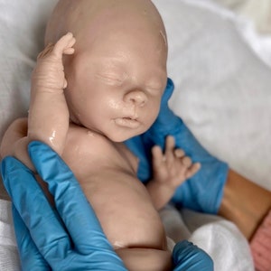 Puede incluir: Un mu&ntilde;eco beb&eacute; realista con los ojos cerrados, que lleva guantes azules, acostado sobre un pa&ntilde;o blanco. El mu&ntilde;eco est&aacute; hecho de silicona y tiene un aspecto realista.