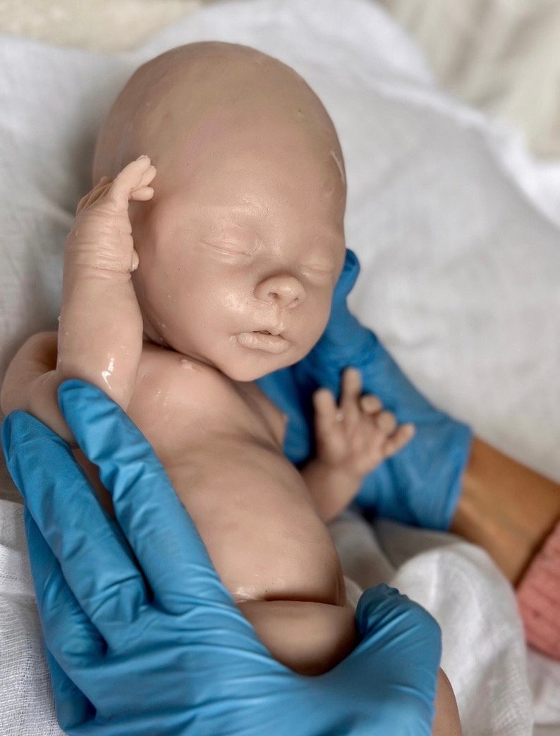 Puede incluir: Un mu&ntilde;eco beb&eacute; realista con los ojos cerrados, que lleva guantes azules, acostado sobre un pa&ntilde;o blanco. El mu&ntilde;eco est&aacute; hecho de silicona y tiene un aspecto realista.