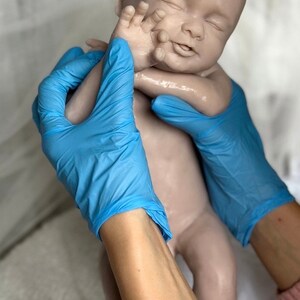 K&ouml;nnte beinhalten: Eine lebensechte Babypuppe in neutralem Farbton mit geschlossenen Augen und einem friedlichen Ausdruck. Die Puppe wird von H&auml;nden mit blauen Handschuhen gehalten. Die Arme des Babys sind leicht angehoben, und die Haut hat eine glatte Textur.