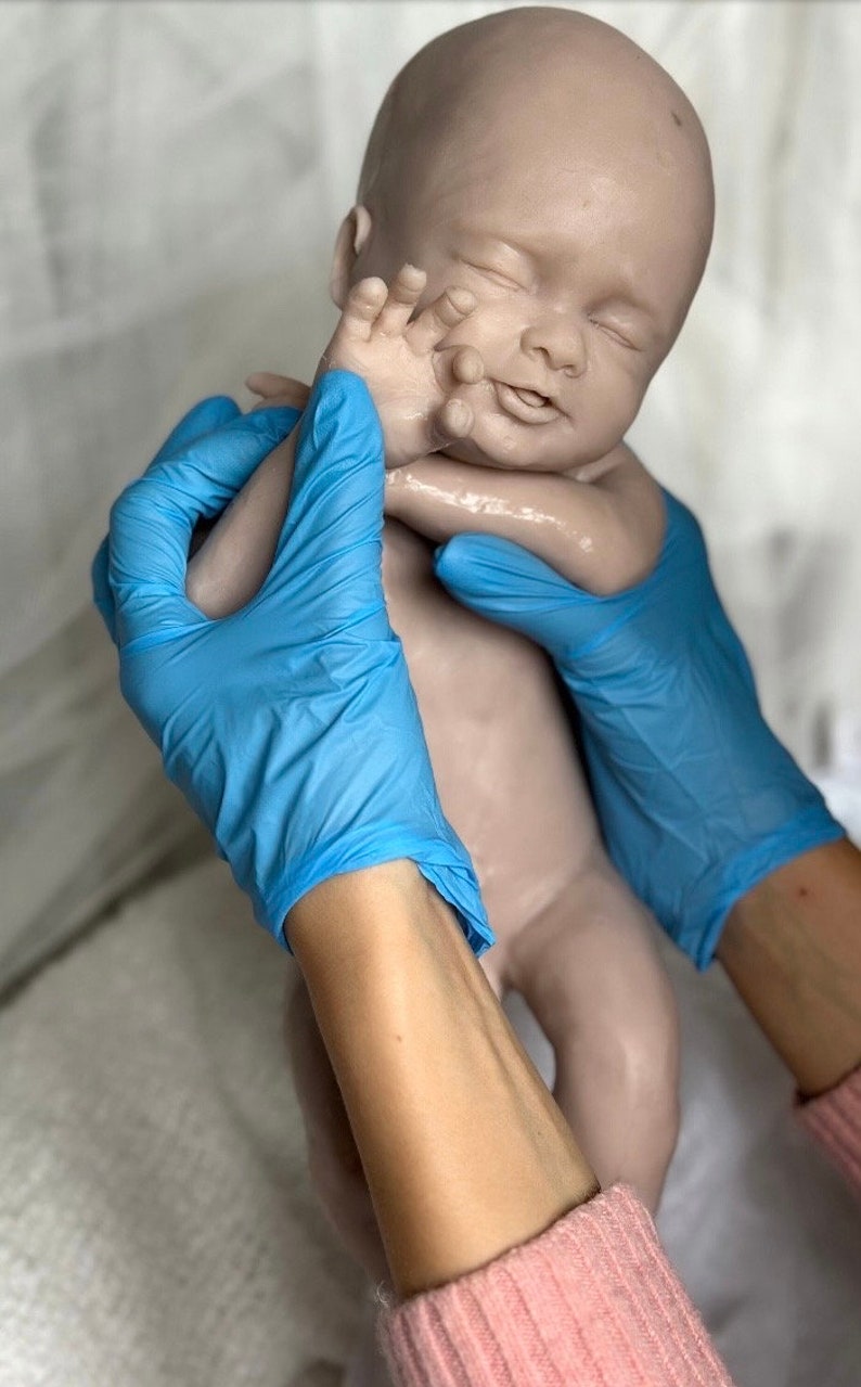 K&ouml;nnte beinhalten: Eine lebensechte Babypuppe in neutralem Farbton mit geschlossenen Augen und einem friedlichen Ausdruck. Die Puppe wird von H&auml;nden mit blauen Handschuhen gehalten. Die Arme des Babys sind leicht angehoben, und die Haut hat eine glatte Textur.