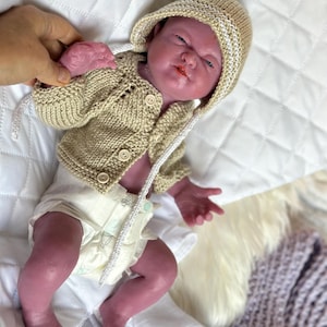 Puede incluir: Una mu&ntilde;eca beb&eacute; realista con una chaqueta con capucha de punto beige y un gorro a juego con ribete blanco. La mu&ntilde;eca tambi&eacute;n lleva un pa&ntilde;al. La mu&ntilde;eca est&aacute; tumbada sobre una superficie acolchada blanca.