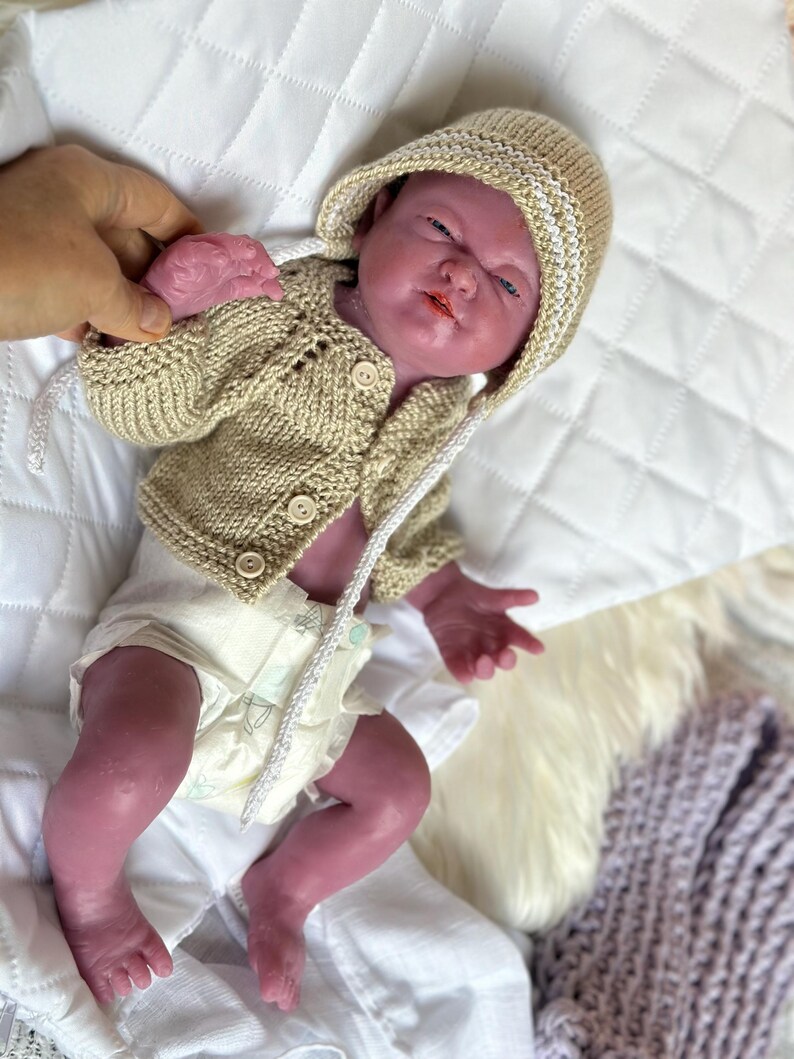 Puede incluir: Una mu&ntilde;eca beb&eacute; realista con una chaqueta con capucha de punto beige y un gorro a juego con ribete blanco. La mu&ntilde;eca tambi&eacute;n lleva un pa&ntilde;al. La mu&ntilde;eca est&aacute; tumbada sobre una superficie acolchada blanca.