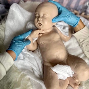 May include: A realistic baby doll with closed eyes and an open mouth, lying on a white blanket. The doll is made of silicone and has a lifelike appearance.