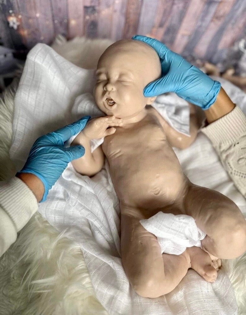May include: A realistic baby doll with closed eyes and an open mouth, lying on a white blanket. The doll is made of silicone and has a lifelike appearance.